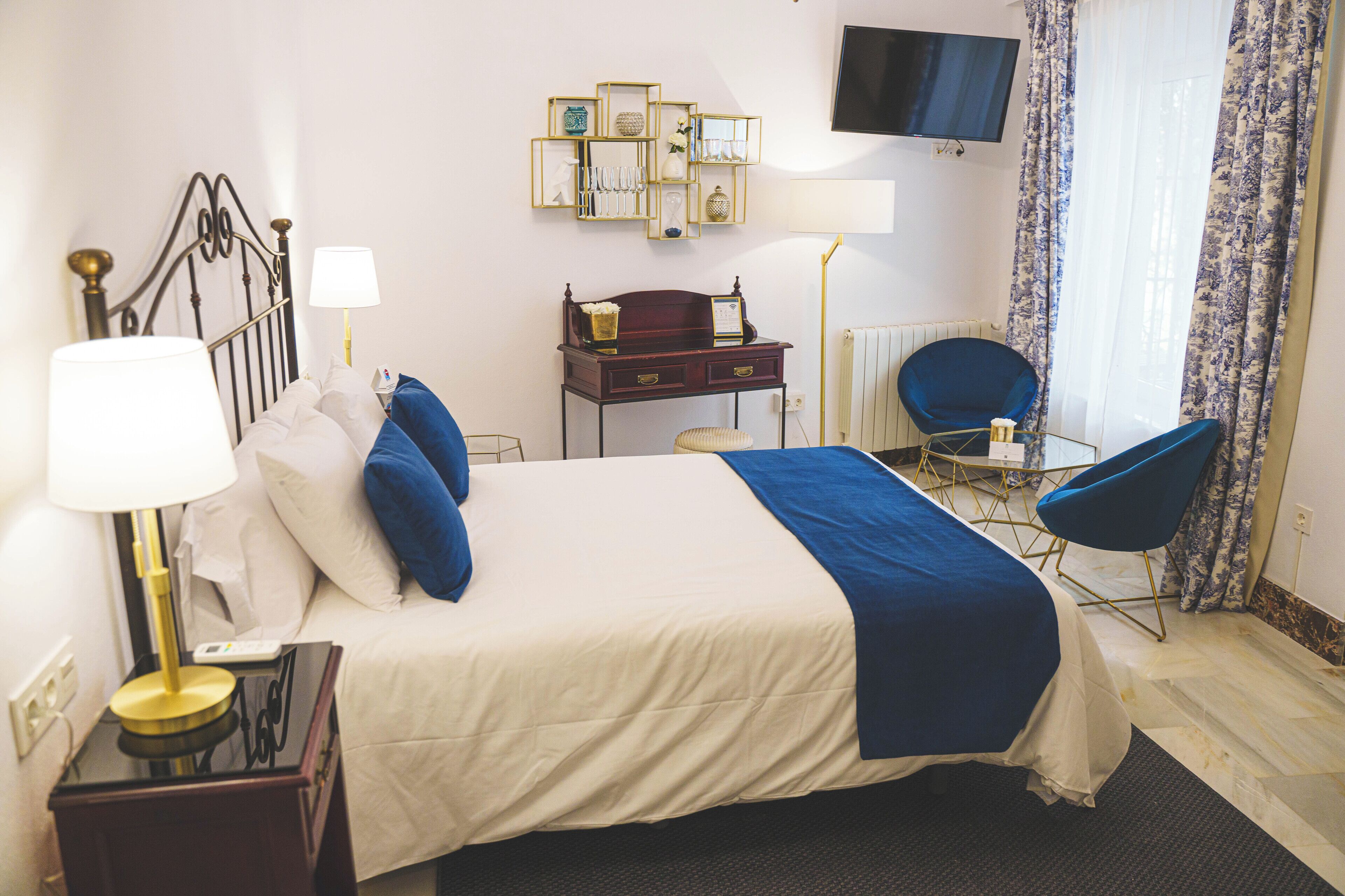 Habitación de hotel con cama, mesita de noche, lámpara, escritorio, silla y televisor en la pared.