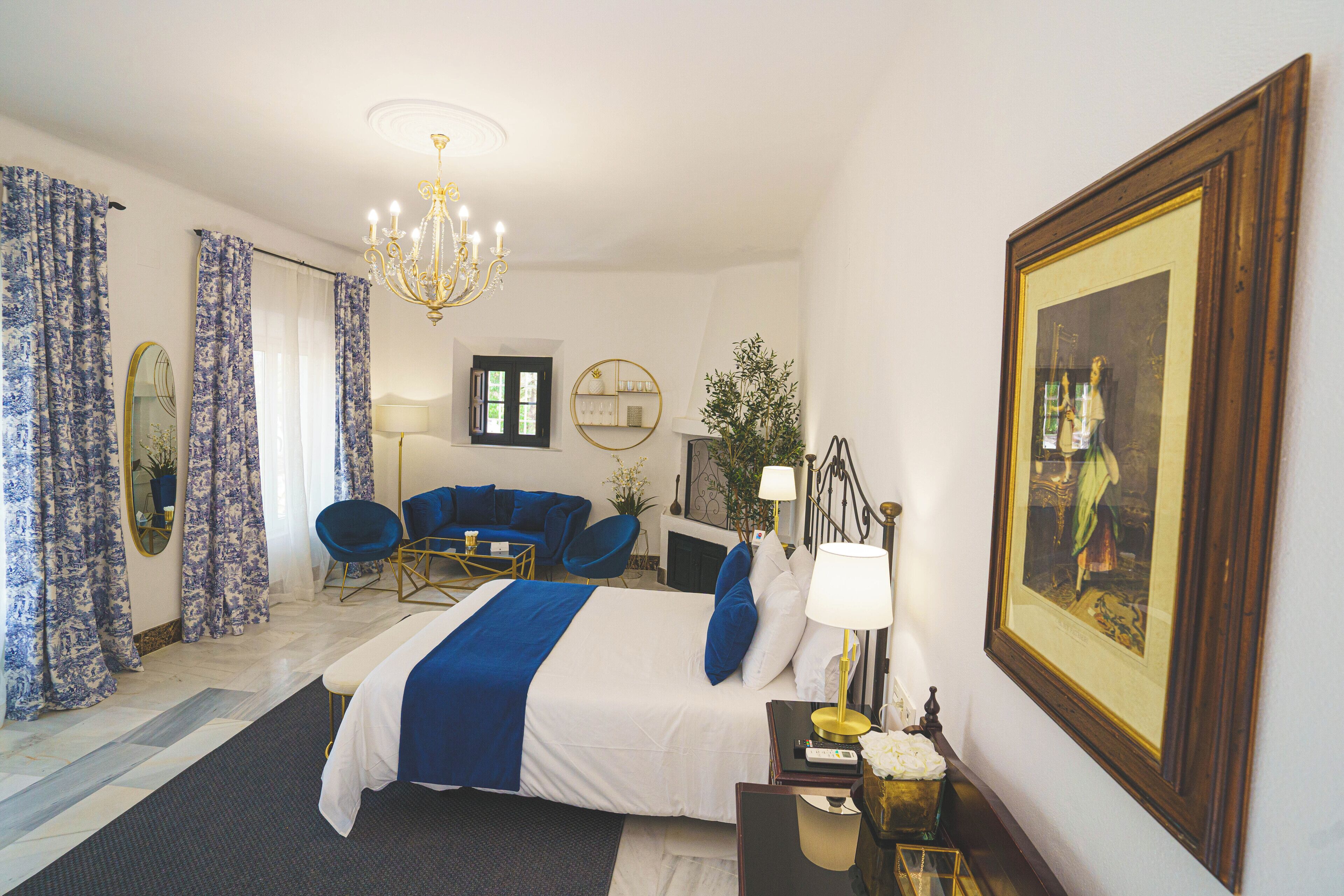 Habitación de hotel con cama, escritorio, silla, sofá, candelabro, cuadro y espejo grande.
