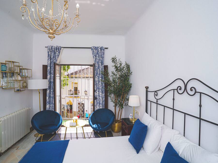 A hotel room with a bed, two chairs, a table, a chandelier, a radiator, and a view of the outside.