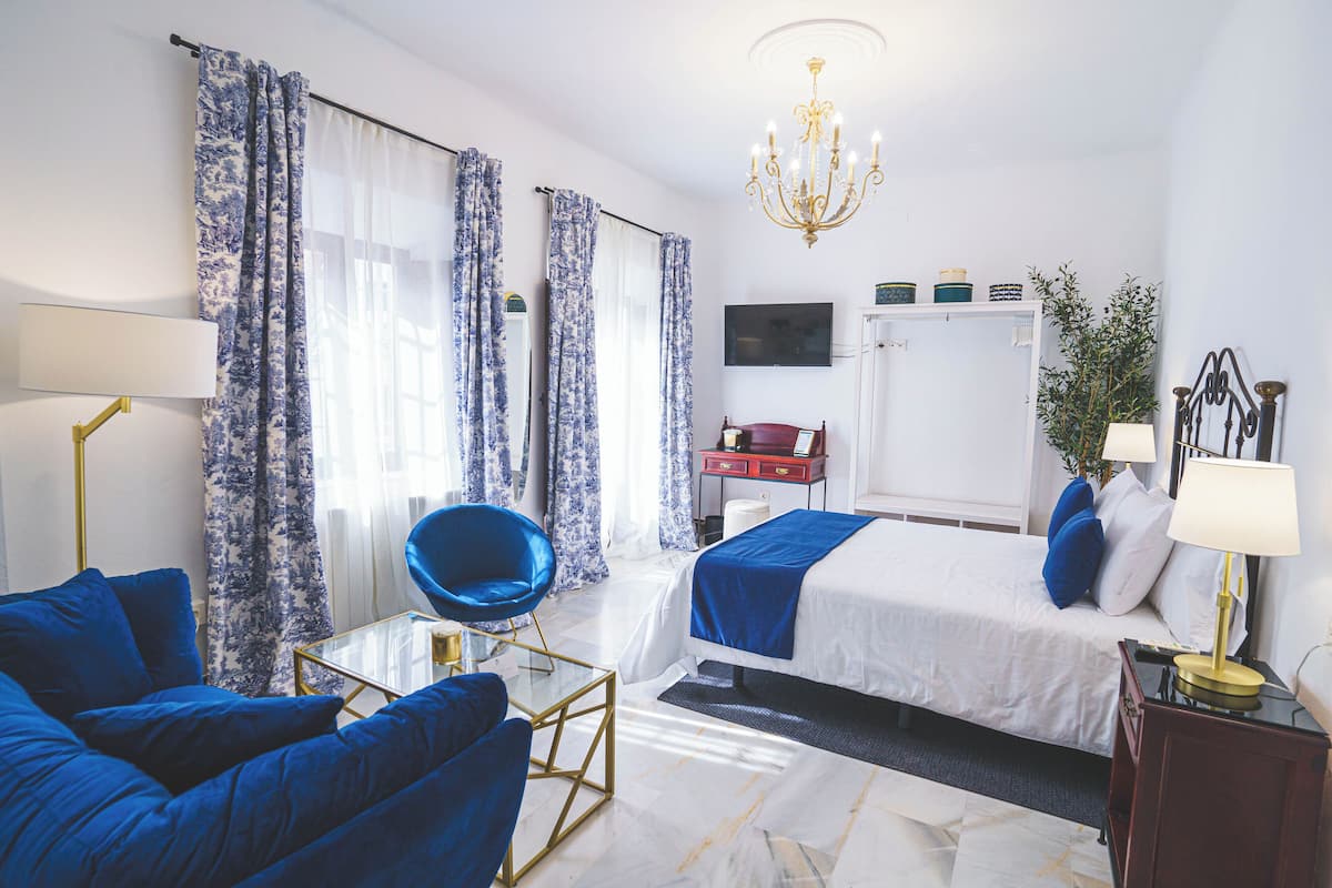 A hotel room with a bed, two blue chairs, a desk, a TV, and a chandelier.