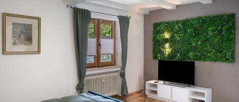 A bedroom with a bed, a TV on a white stand, a window with curtains, and a green wall decoration.