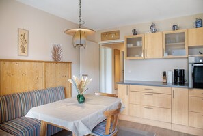 A dining area with a table, chairs, and a vase of dried plants. There is a wooden bench with striped cushions, a hanging light fixture, and wooden cabinets with glass doors.