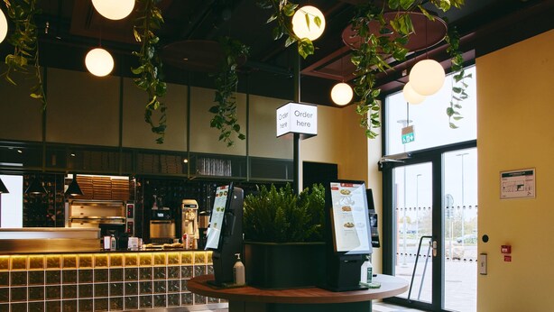 Un interior moderno con suelo de cuadros, una recepción, un cartel de menú y plantas colgantes.