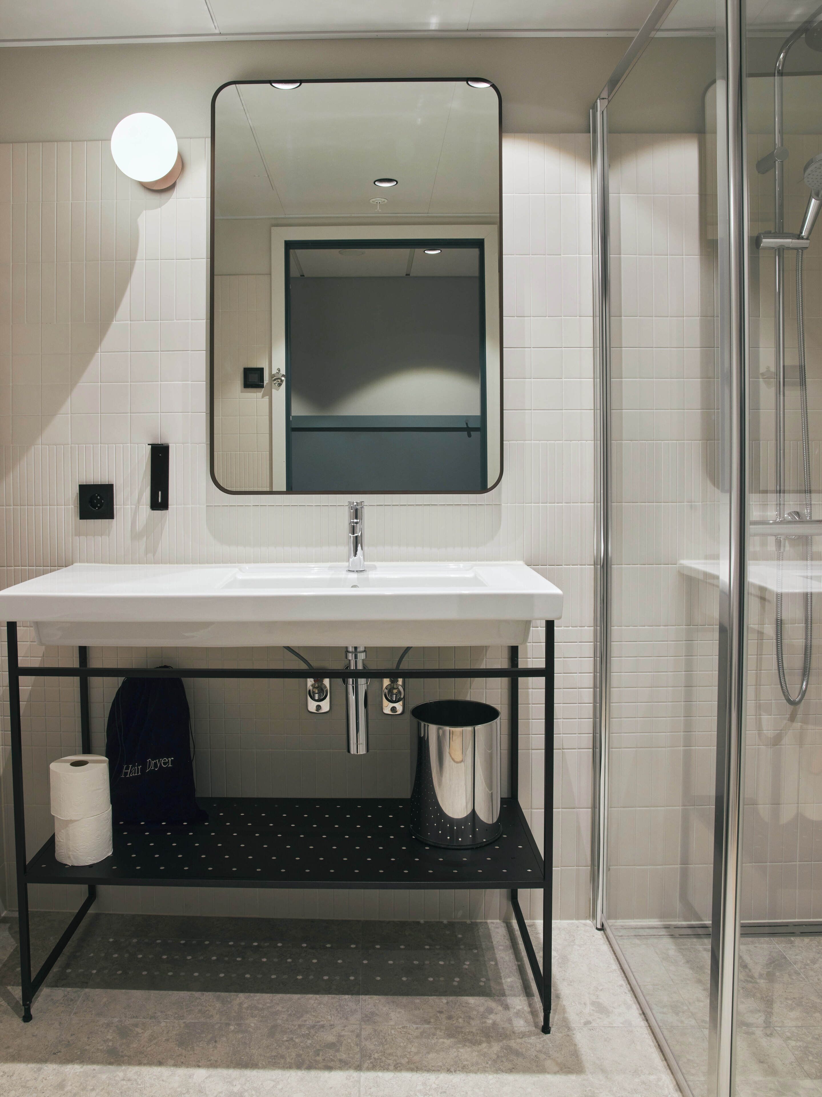 A modern bathroom with a white sink, a mirror, a glass shower enclosure, and a black shelf with a hair dryer and toilet paper.