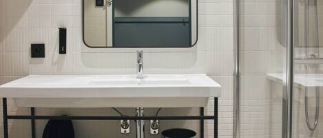 A modern bathroom with a white sink, a mirror, a glass shower enclosure, and a black shelf with a hair dryer and toilet paper.