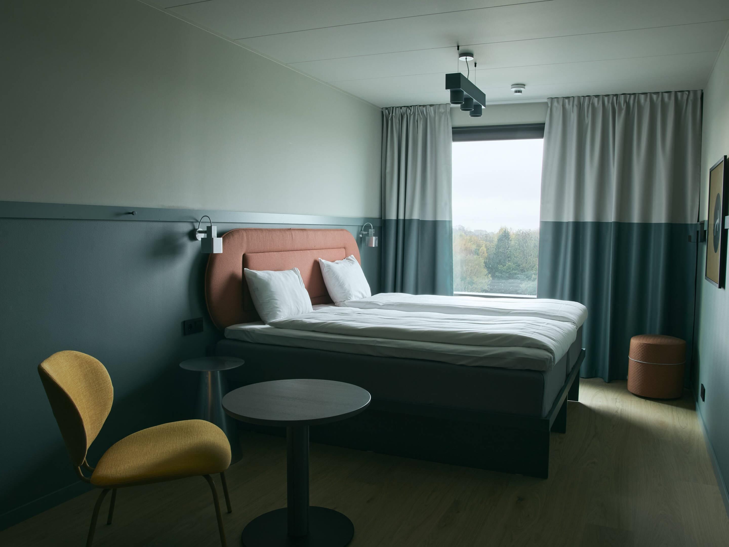 Una camera d'albergo con un letto, una sedia, un tavolino e una finestra con tende.