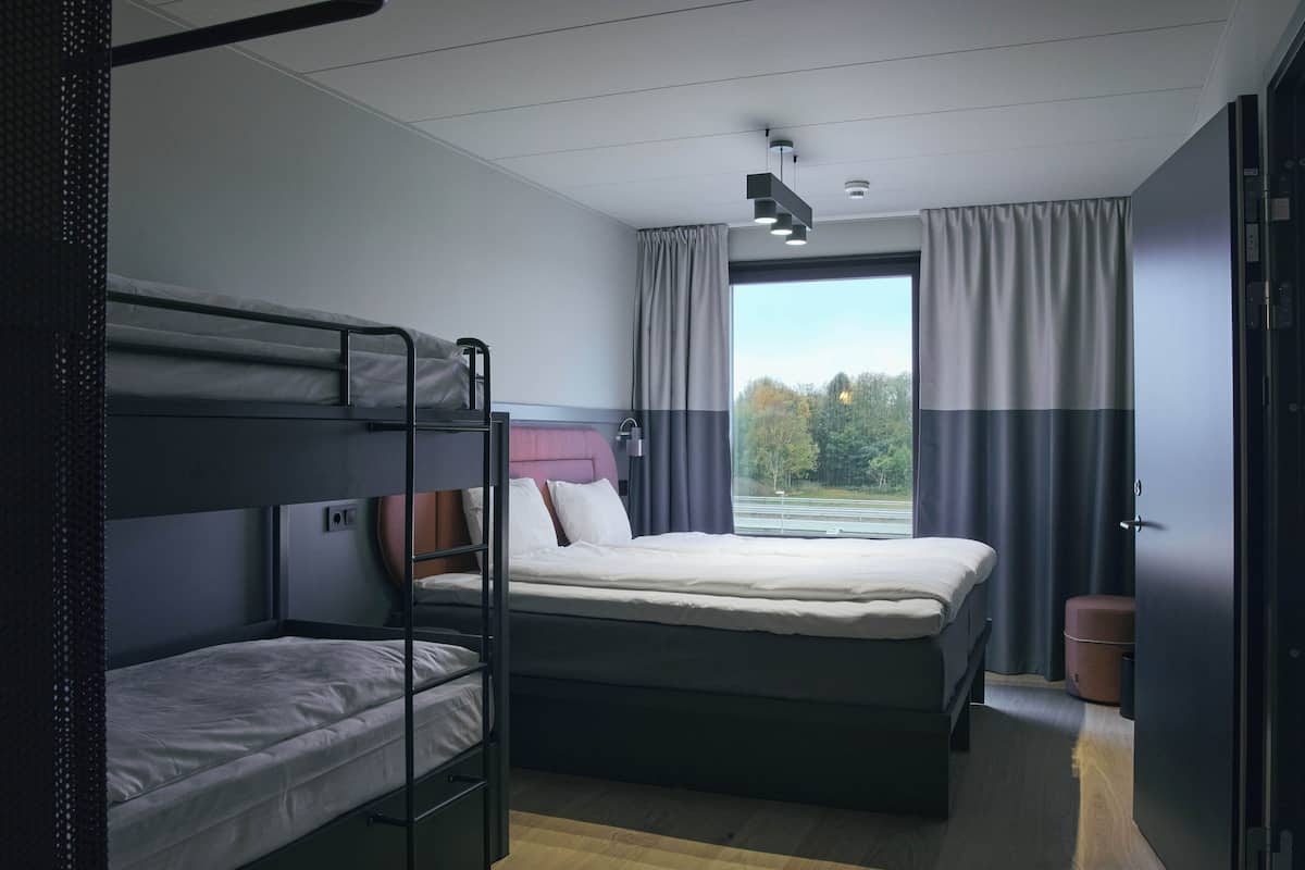 A modern hotel room with bunk beds, a large window with curtains, a wooden floor, and a door with a handle.
