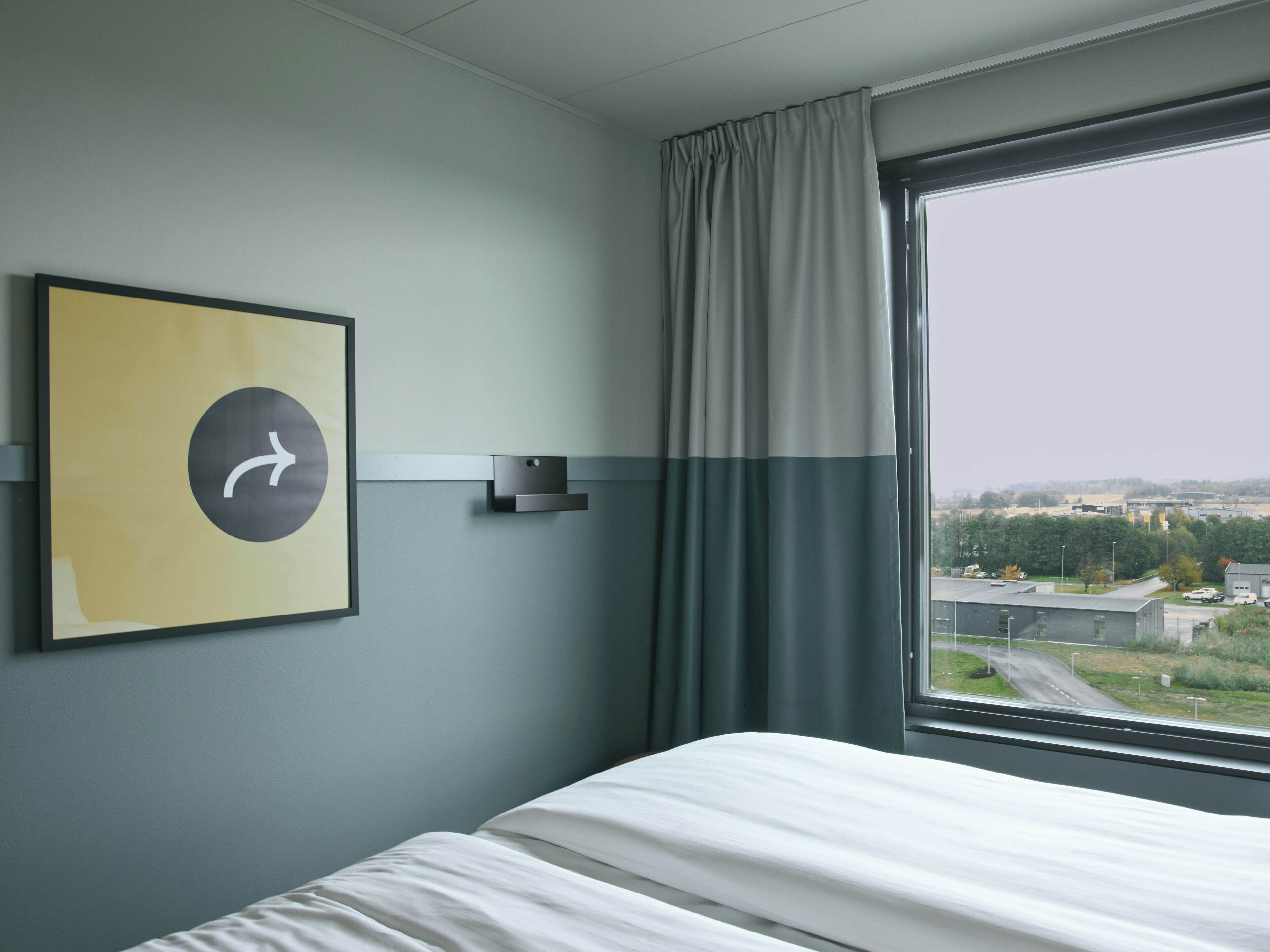 A hotel room with a bed, a wall-mounted clock, a window with curtains, and a view of the outside.