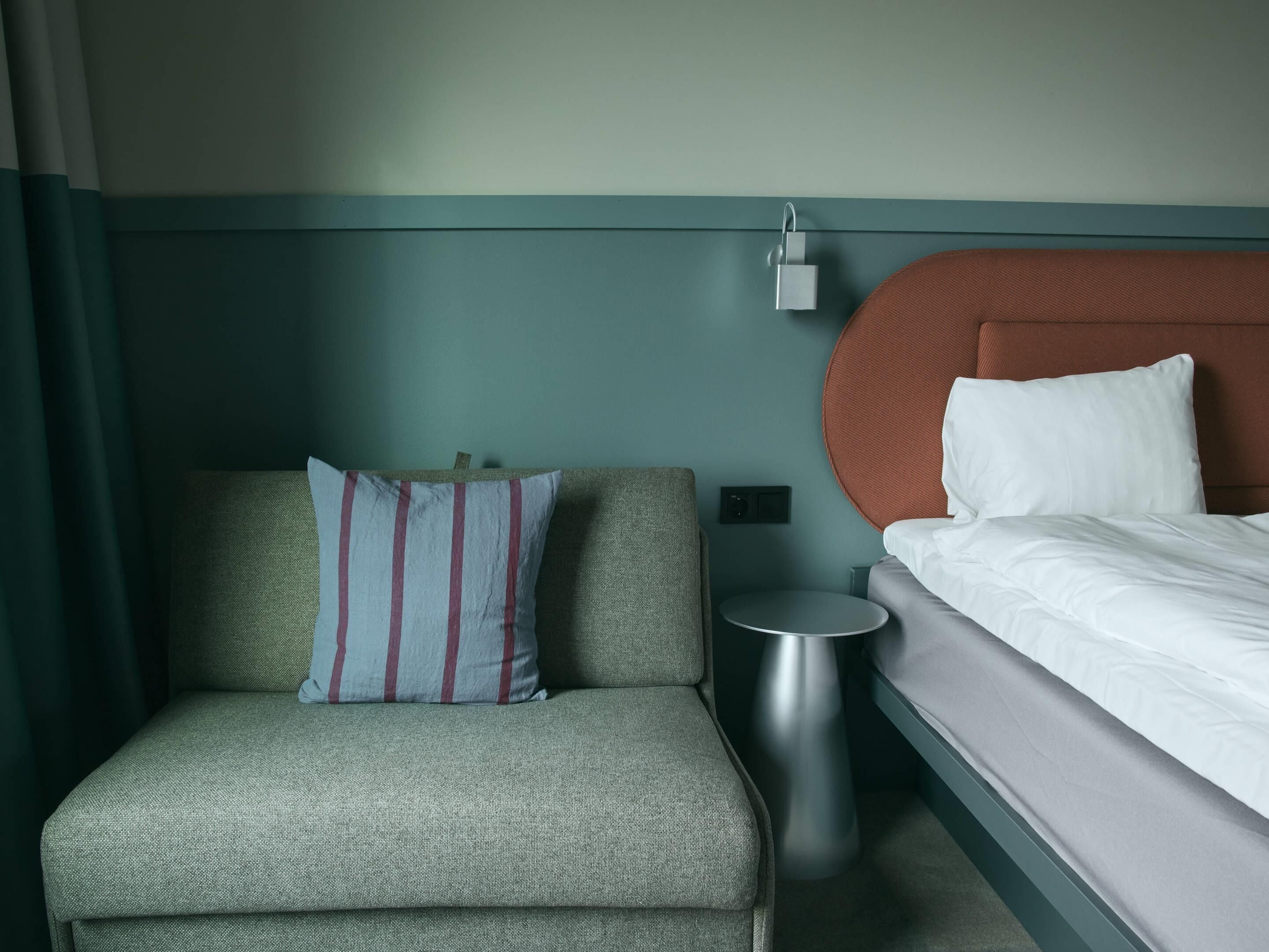 A hotel room with a bed, a small round table, a sofa with a striped pillow, and a wall-mounted charging station.