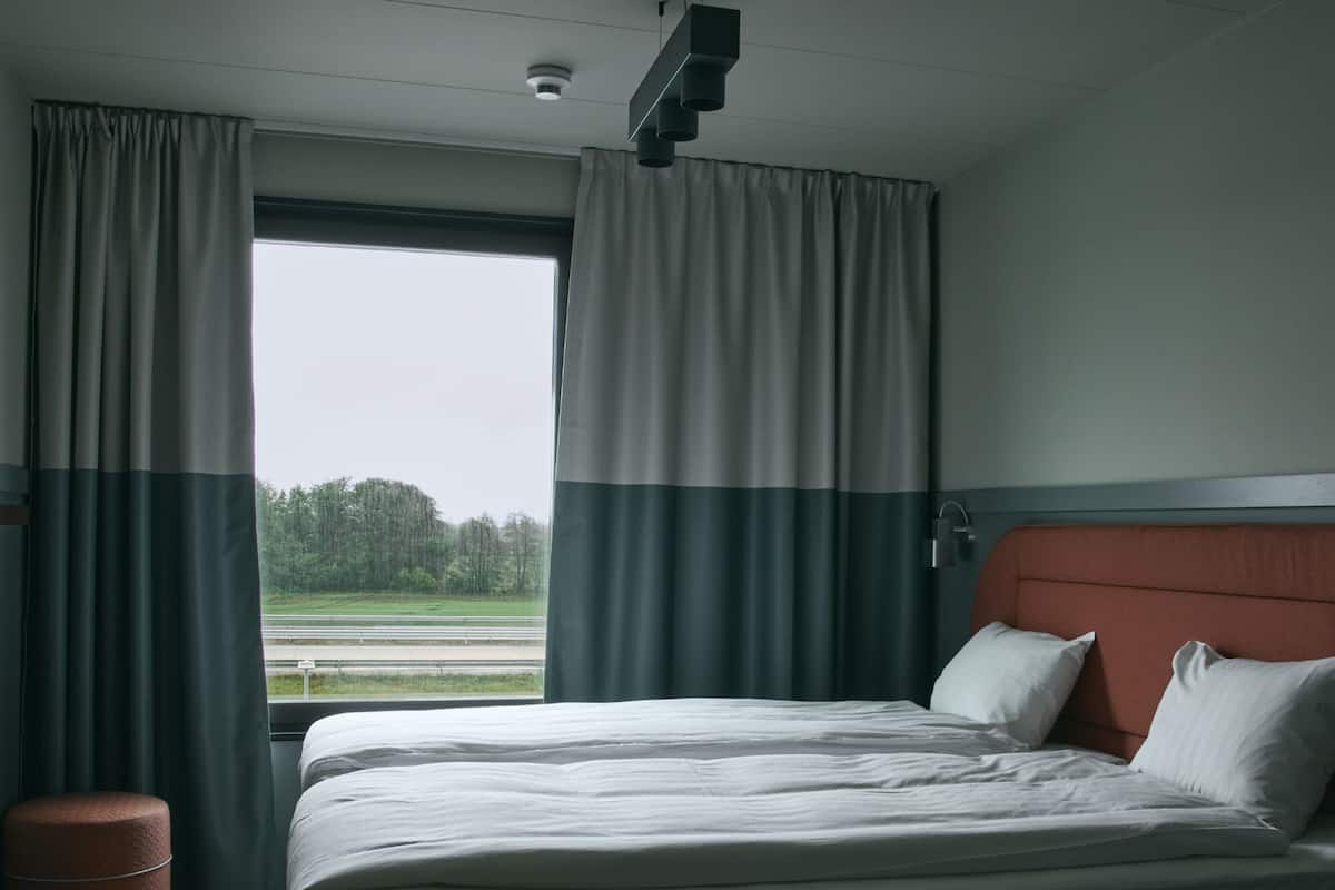 A hotel room with a bed, curtains, a window with a view of a green landscape, and a ceiling light fixture.
