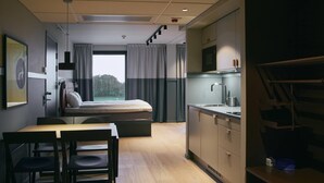 Una habitación de hotel compacta con cama, una kitchenette pequeña, una zona de comedor con mesa y sillas, y un ventanal con cortinas.