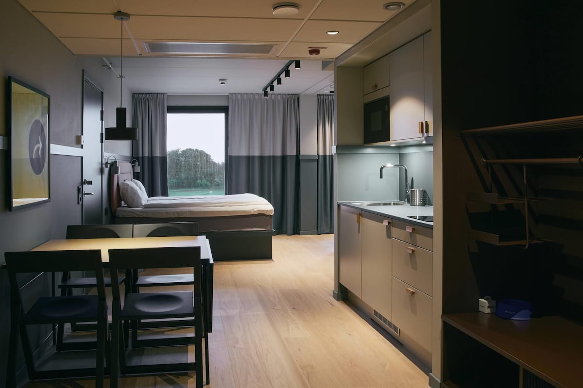 A compact hotel room with a bed, a small kitchenette, a dining area with a table and chairs, and a large window with curtains.