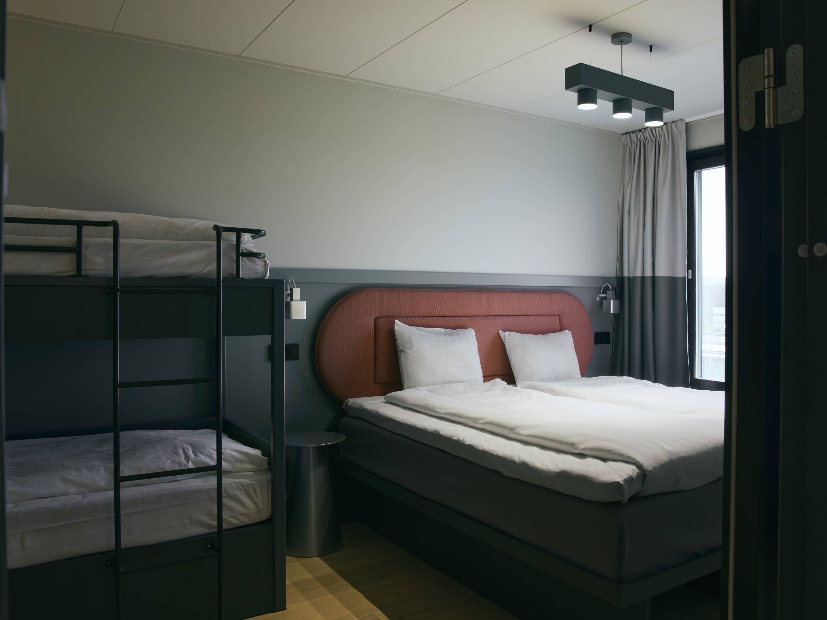 A hotel room with a bunk bed, a single bed, a bedside table, a lamp, and a window with curtains.