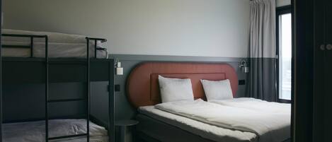 A hotel room with a bunk bed, a single bed, a bedside table, a lamp, and a window with curtains.
