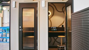 Un vestíbulo de hotel con piso a cuadros, una puerta negra con persianas verticales, un taburete pequeño y una computadora portátil sobre una estantería.
