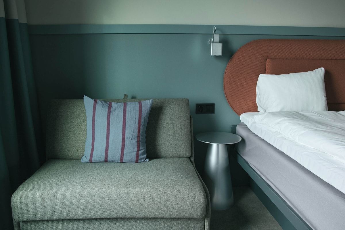 A hotel room with a grey sofa, a small round side table, a bed with white bedding, and a wall-mounted light fixture.