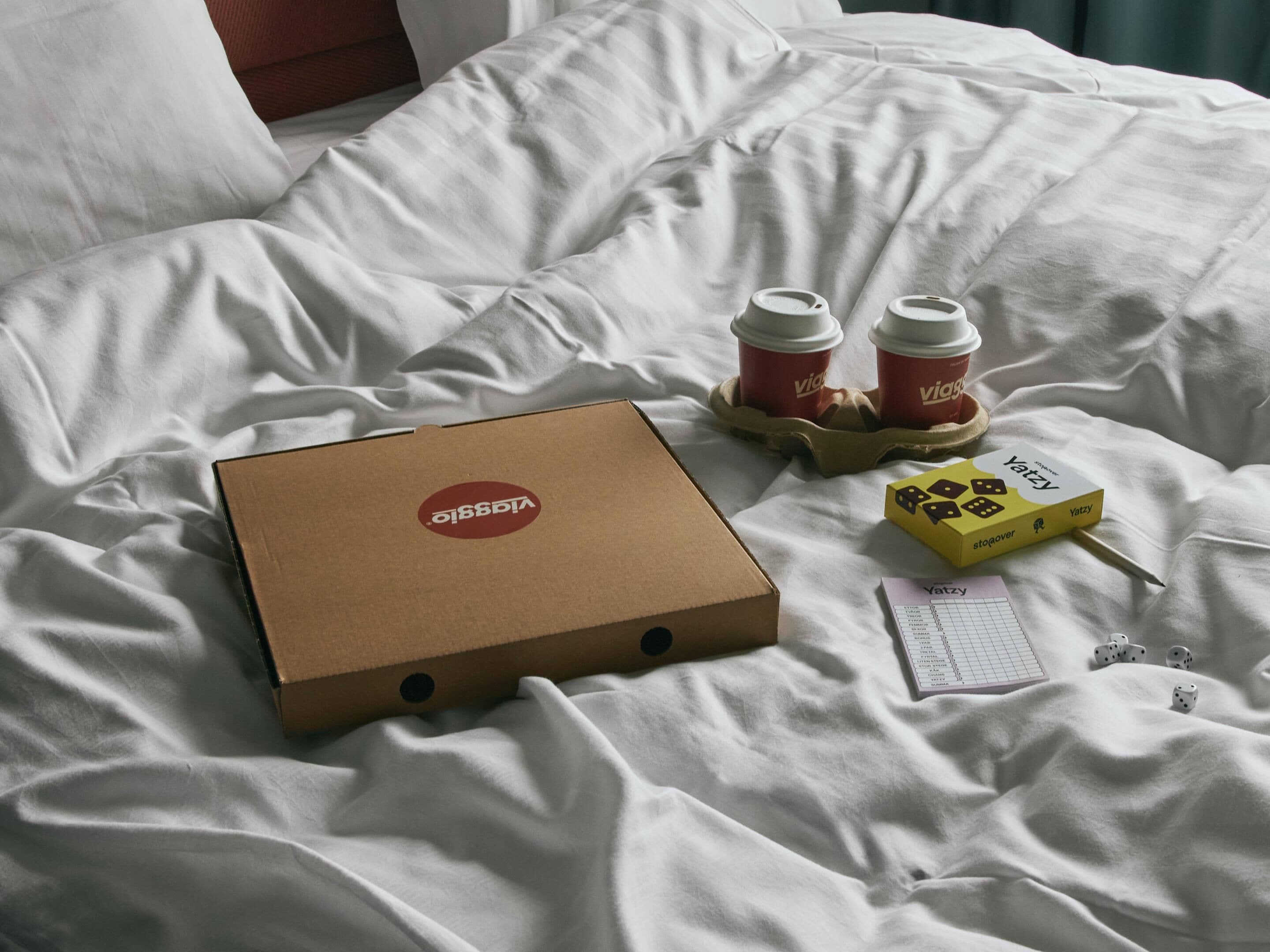 Un lit avec une couette blanche, une boîte en carton ornée d’un logo, deux gobelets à café jetables, une boîte jaune contenant des dés et un reçu.