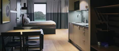 A compact hotel room with a bed, a small kitchenette, a dining area with a table and chairs, and a large window offering a view of the outdoors.