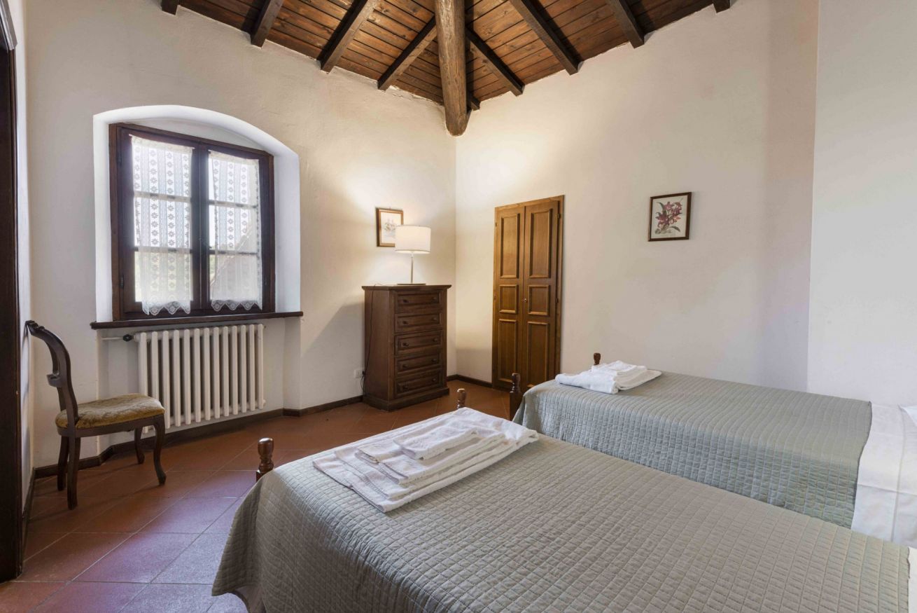 A bedroom with two beds, a wooden dresser, a chair, a window with patterned curtains, and a radiator.