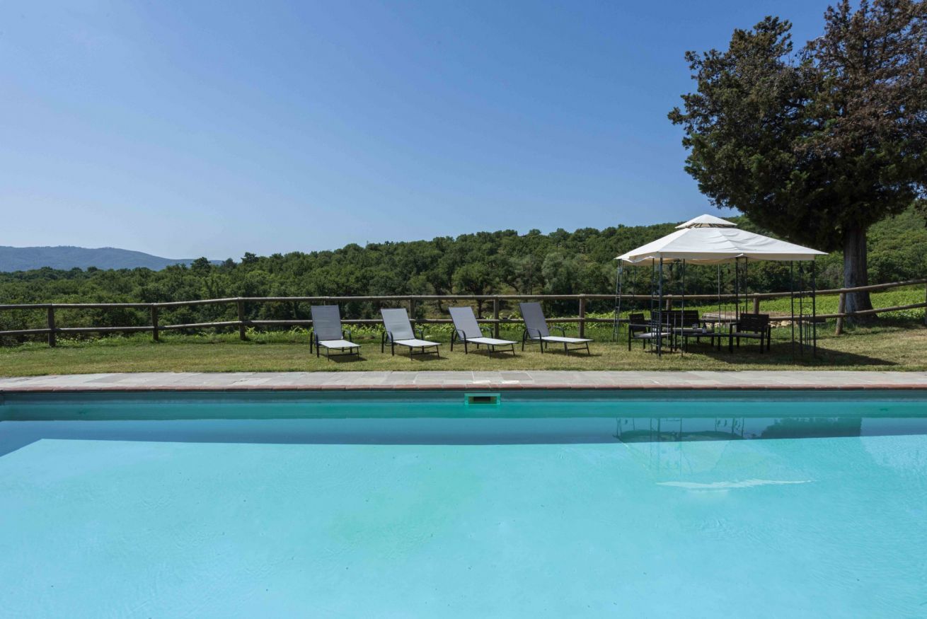 Une piscine entourée de chaises longues et d’une table placée sous un auvent, le tout bordé d’une pelouse et d’arbres.