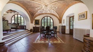 A spacious room with a high arched ceiling, a dining table with chairs, a patterned rug, and large windows.