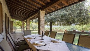 A covered outdoor dining area with a long table set for four, stone pillars, and a view of trees and greenery.