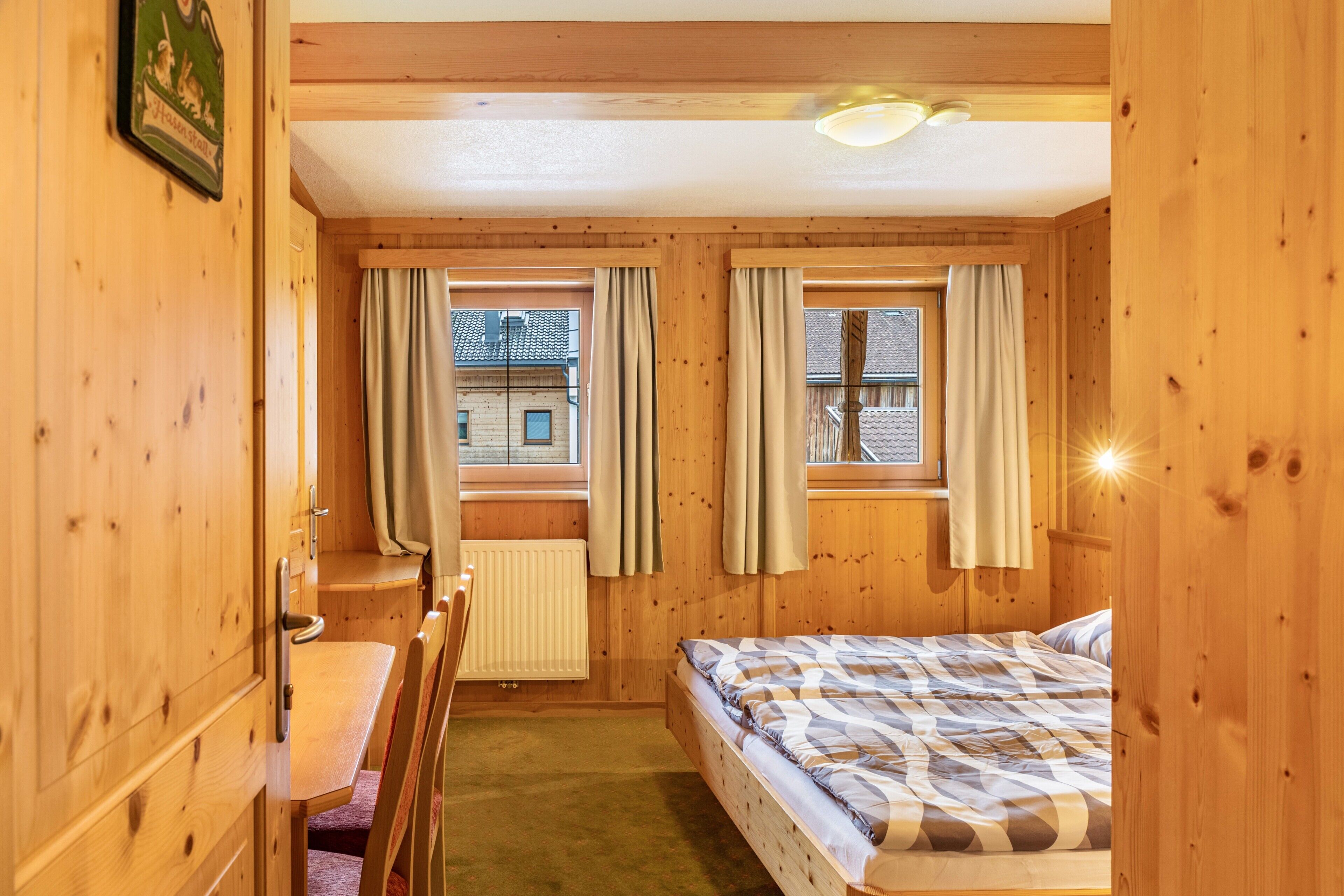 Ein Zimmer mit Holzwänden, einem Bett, einem Schreibtisch und zwei mit Vorhängen versehenen Fenstern.