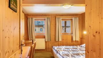 Ein Zimmer mit Holzwänden, einem Bett, einem Schreibtisch und zwei mit Vorhängen versehenen Fenstern.