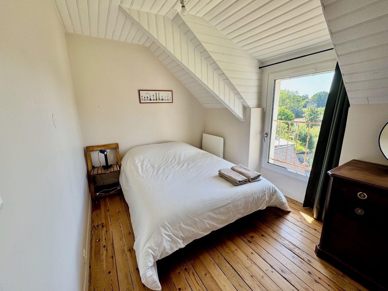 A bedroom with a bed, wooden floor, a small bedside table, a nightstand, a dresser, a window with a view, and a framed picture on the wall.
