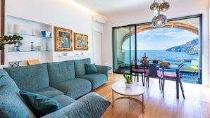 Una sala de estar moderna con un sofá grande, una mesa de comedor con sillas, una mesita redonda con una planta y vistas al mar a través de un ventanal.