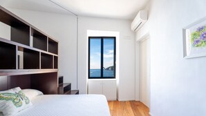 Un dormitorio con cama, ventana con vista al mar, estante empotrado y puerta blanca.