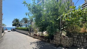 Une rue pavée, bordée d’un côté par un mur de pierre et de verdure, un ciel bleu dégagé et, au fond, un bâtiment doté d’un balcon.
