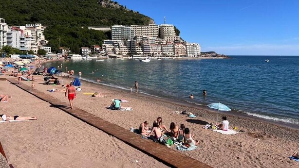 Une plage avec une promenade en bois, des gens qui prennent des bains de soleil et, au loin, un grand bâtiment d’hôtel.