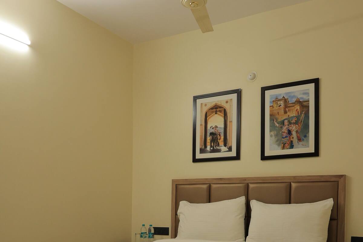 A neatly made bed in a hotel room with a ceiling fan, two framed pictures on the wall, and a bedside table with a water bottle.