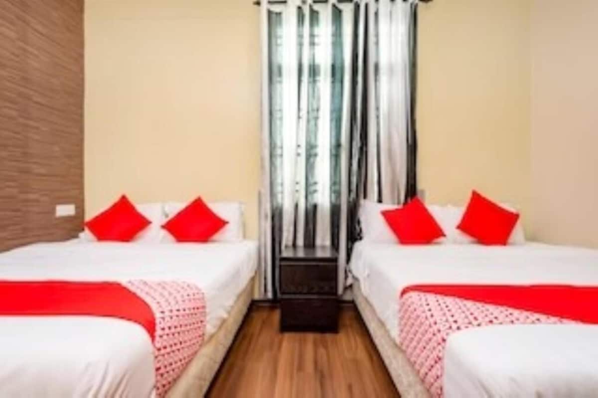 A hotel room with two beds, each with red pillows and a red blanket. There is a window with white curtains and a view of the outside.