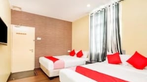 Camera d'albergo con due letti, ciascuno con cuscini rossi e copripiumone bianco. Si nota una porta a sinistra e una finestra con tende.