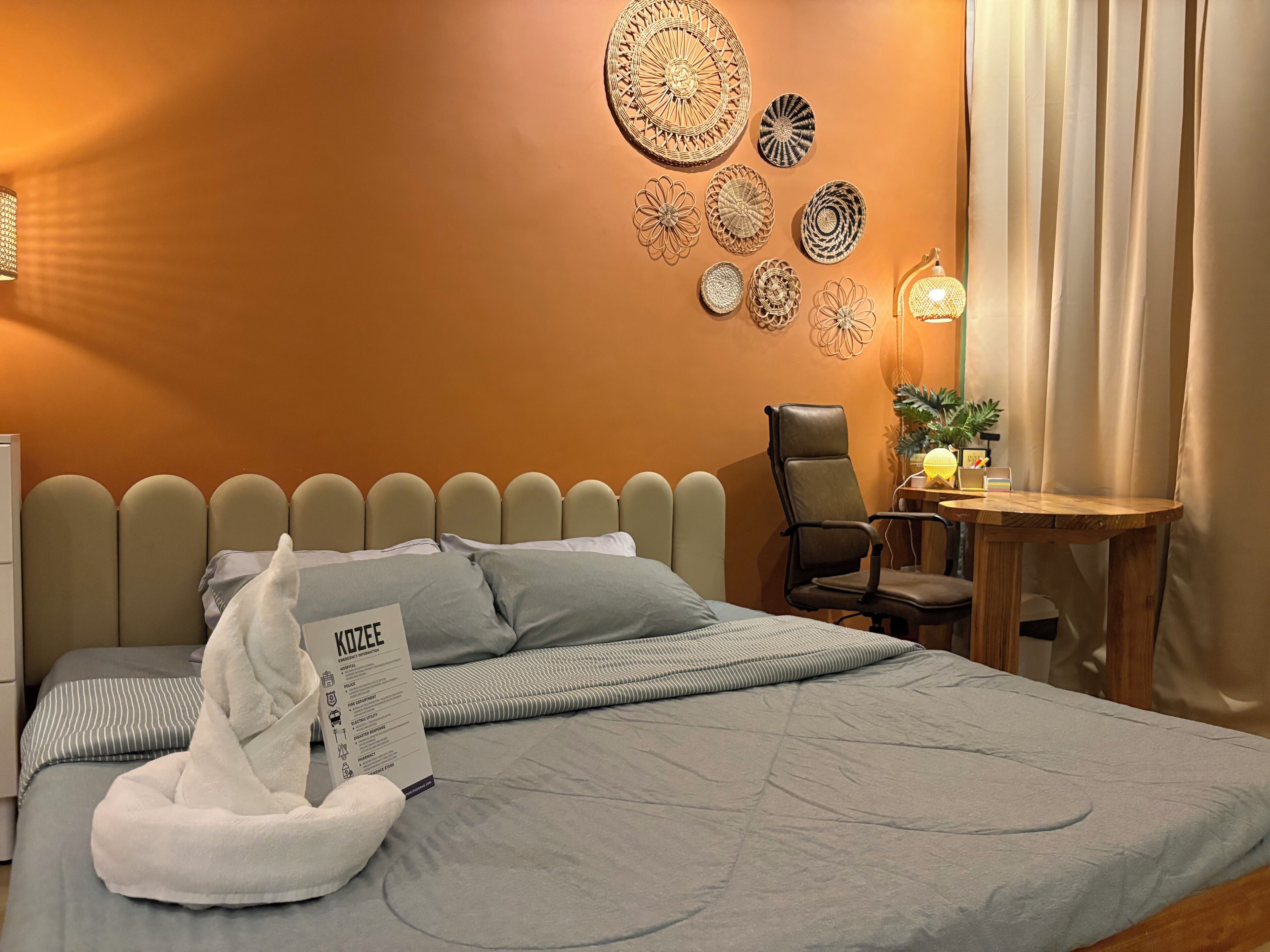 Una cama bien hecha con una almohada y una caja de pañuelos, una silla, una mesita con una lámpara y una decoración artística en la pared.
