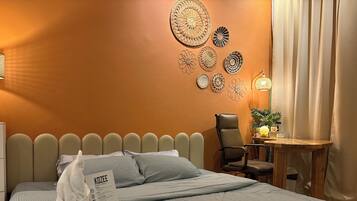 Una cama bien hecha con una almohada y una caja de pañuelos, una silla, una mesita con una lámpara y una decoración artística en la pared.