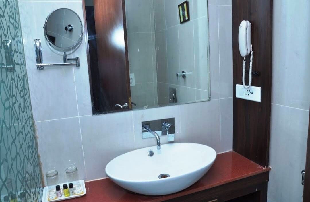 A bathroom with a white sink, a mirror, a handheld mirror, a telephone, and a glass shower enclosure.