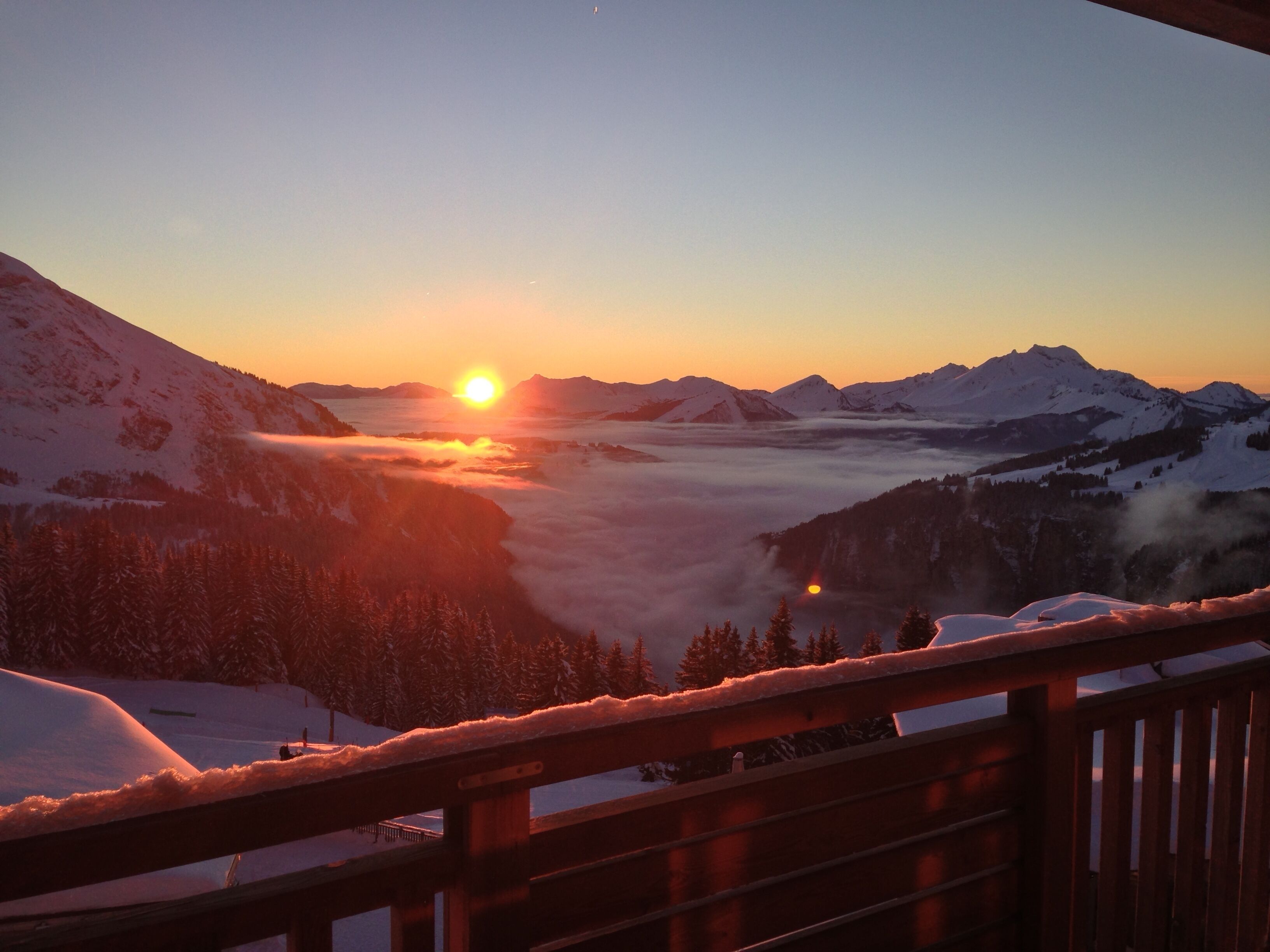 A mountain landscape with a sunrise, snow-covered peaks, and a wooden railing in the foreground.
