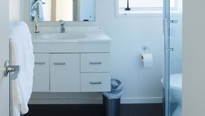 Ein modernes Badezimmer mit einem weißen Waschtisch, einem großen Spiegel, einer Duschkabine aus Glas und einem Fenster mit Jalousien.