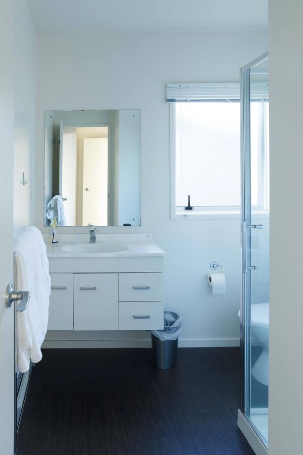 Un baño moderno con un tocador blanco, un espejo grande, una cabina de ducha de vidrio y una ventana con persianas.