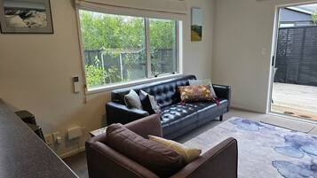 A modern living room with a black leather sofa, a brown armchair, and a large window offering a view of a backyard.
