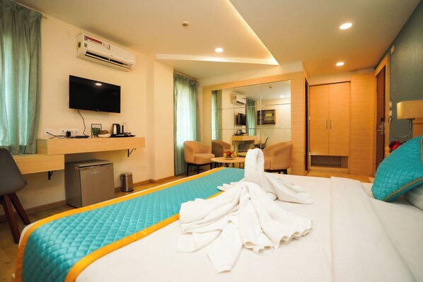 A hotel room with a bed, a desk, a television, an air conditioner, a small fridge, and a bathroom visible through a glass partition. - Santrina Hotels (Bengaluru)