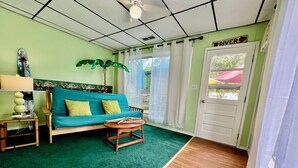 Quarto com sofá verde, uma pequena mesa de madeira, tapete verde e uma porta com a inscrição 'RIVER'.