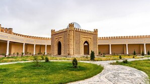 Une cour intérieure abritant une structure centrale surmontée d’un dôme et dotée d’entrées en arc de cercle, le tout entouré d’un mur bas agrémenté de colonnes et d’un chemin pavé de pierres.