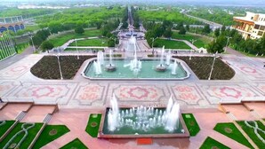 Une grande fontaine richement décorée, dotée de nombreux jets d’eau, se trouve au centre d’une vaste place, entourée de pelouses bien entretenues et de sentiers.