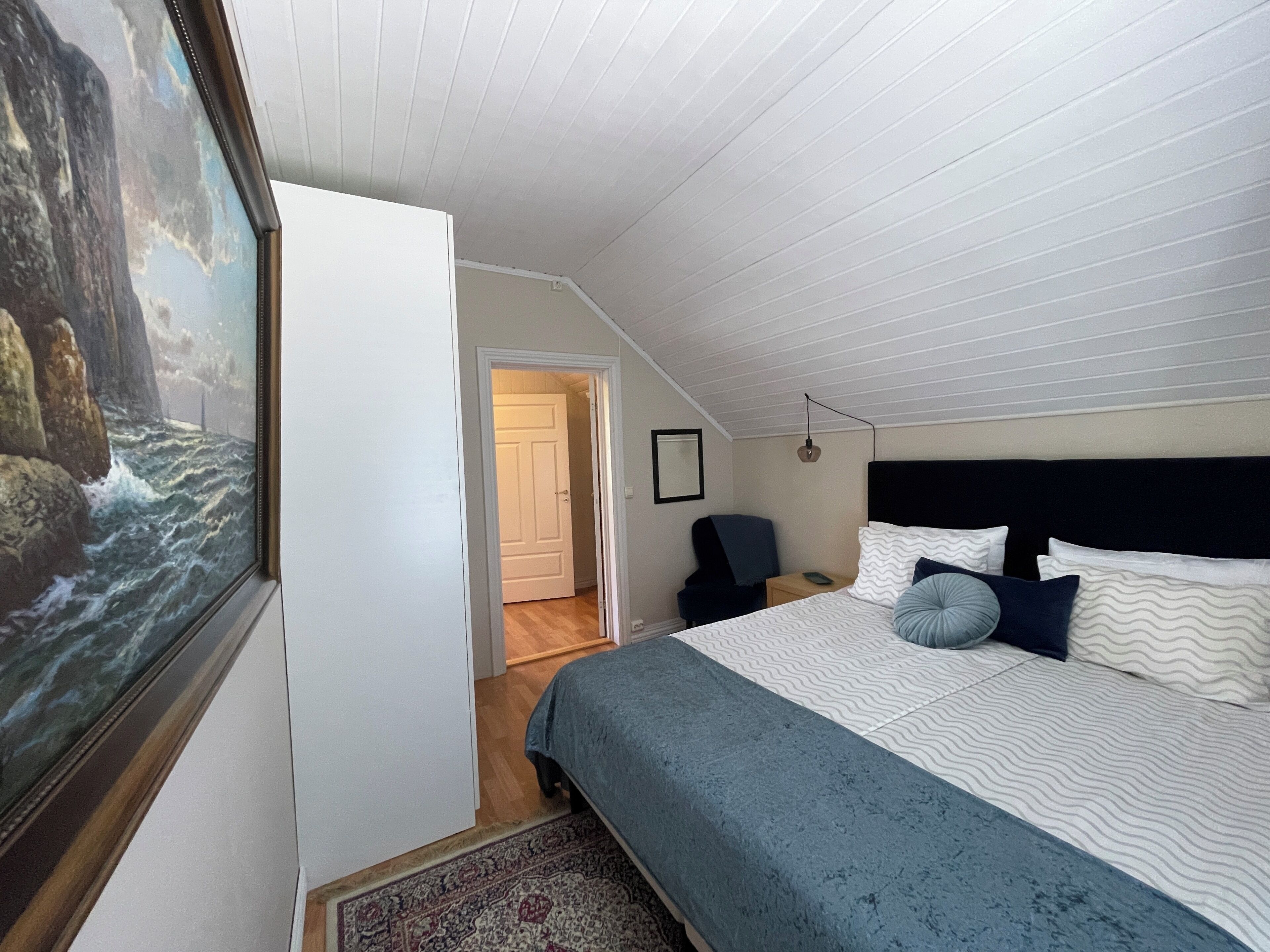 A bedroom with a bed, a chair, a painting, and a door leading to another room.