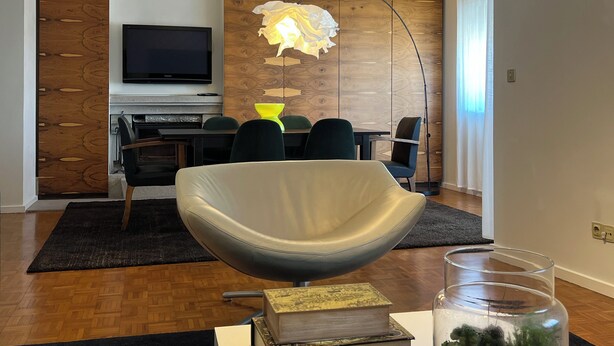 Een moderne woonkamer met een grote, gebogen witte fauteuil, een flatscreen televisie aan een houten wand bevestigd, een eethoek met een tafel en stoelen, en een decoratieve lamp.