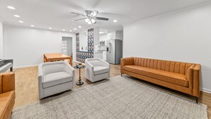 Een moderne woonkamer met een piano, twee fauteuils, een bank, een eettafel en een keuken op de achtergrond.